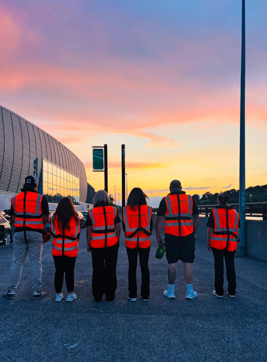 Six Re-A-Suir volunteers in hi-vis vests standing side by side on the Waterford Greenway under a pink and orange sunset
