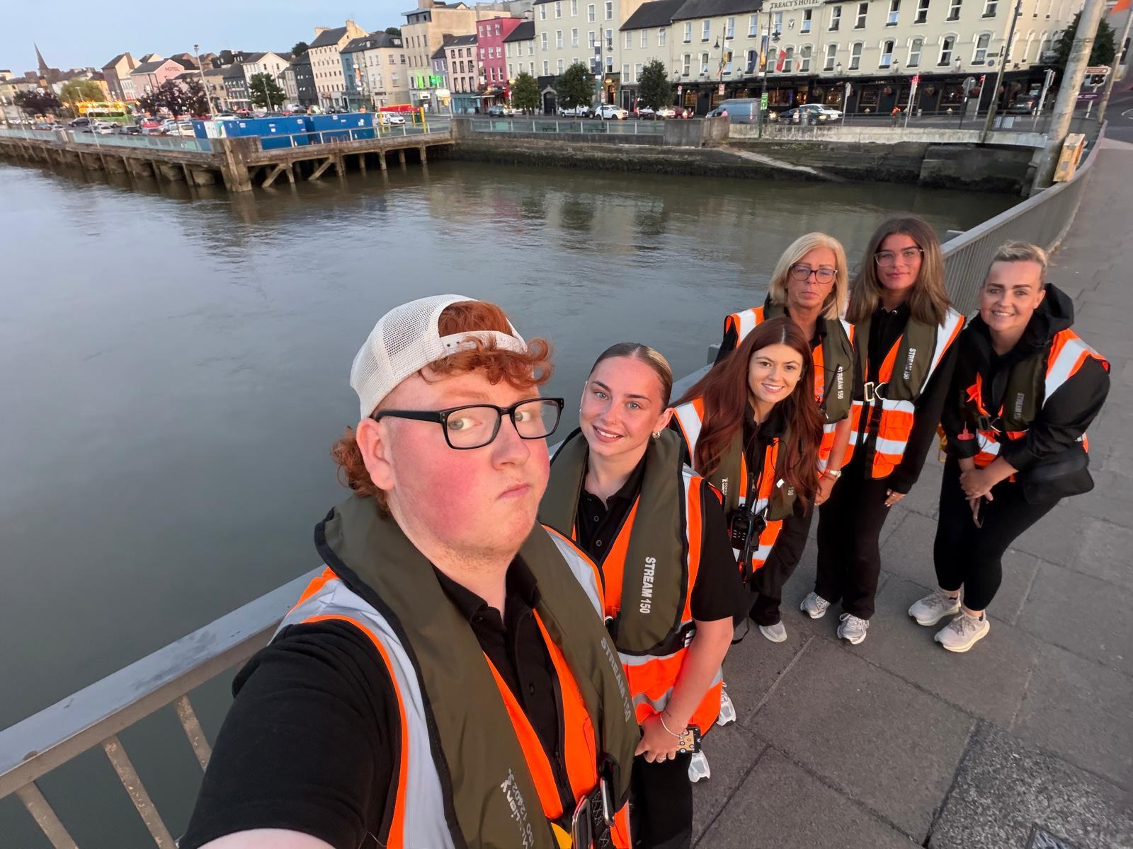 Six smiling Re-A-Suir volunteers in hi-vis vests and lifejackets taking a selfie on the bridge, with the quays behind them