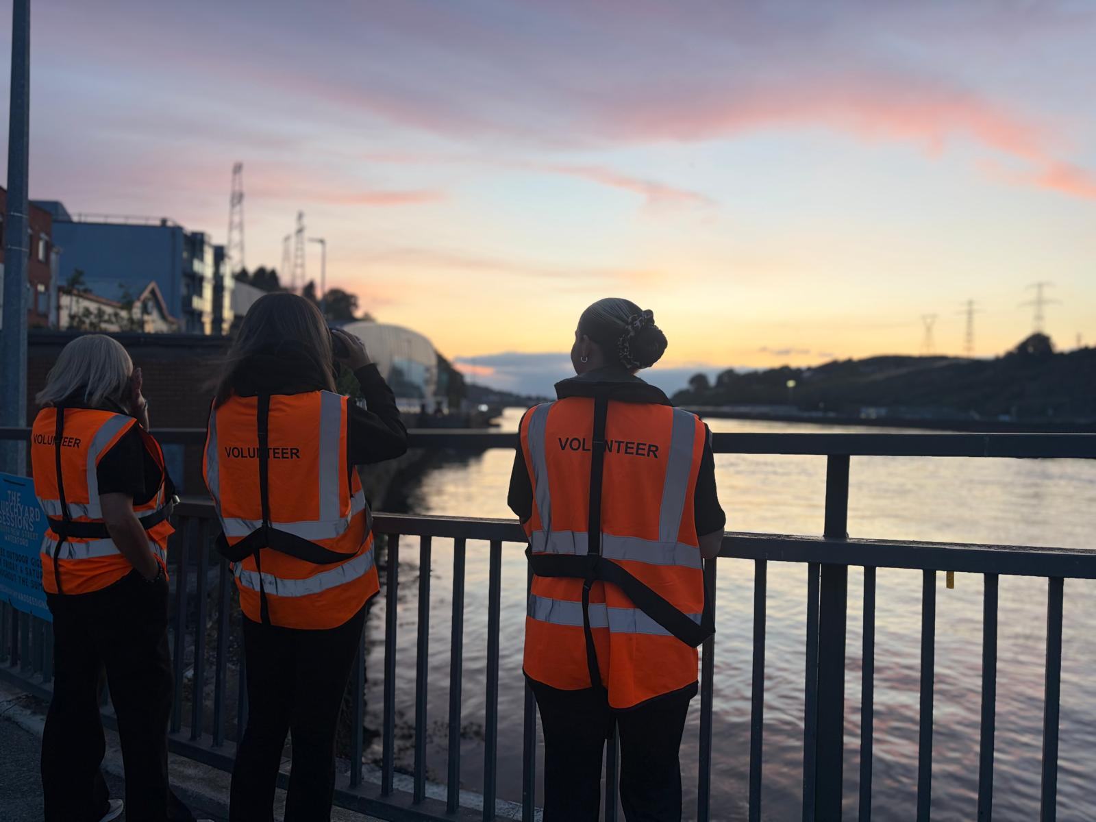 Three volunteers in hi-vis vests at the bridge railing, watching the River Suir at sunset