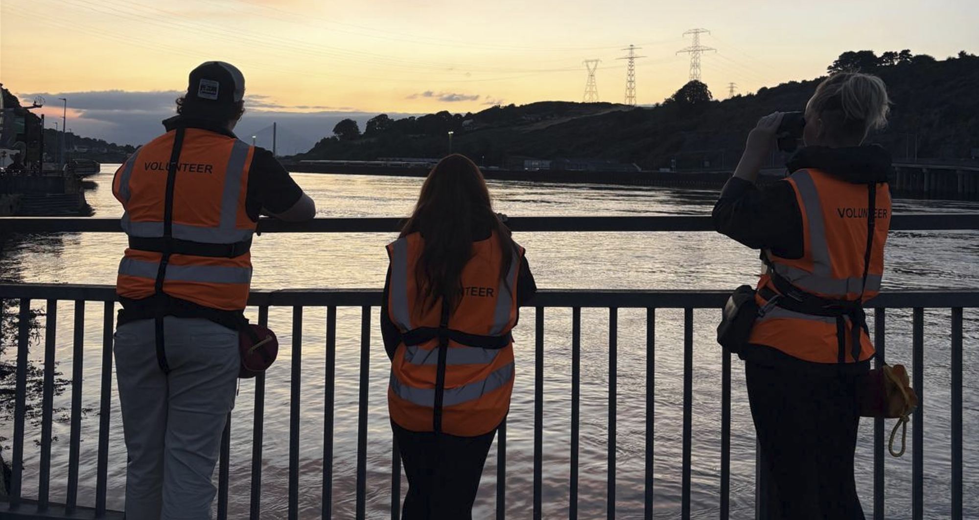 Three volunteers at the bridge railing watching the River Suir at sunset