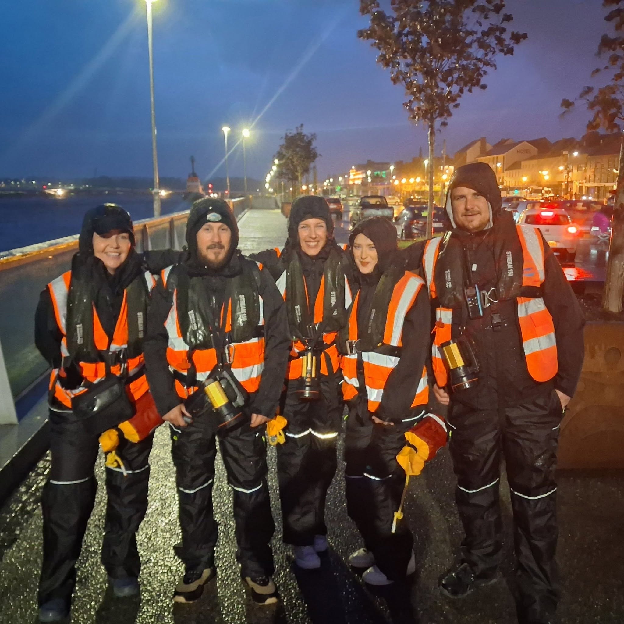 Five volunteers in orange rain gear with torches on a wet quayside at dusk